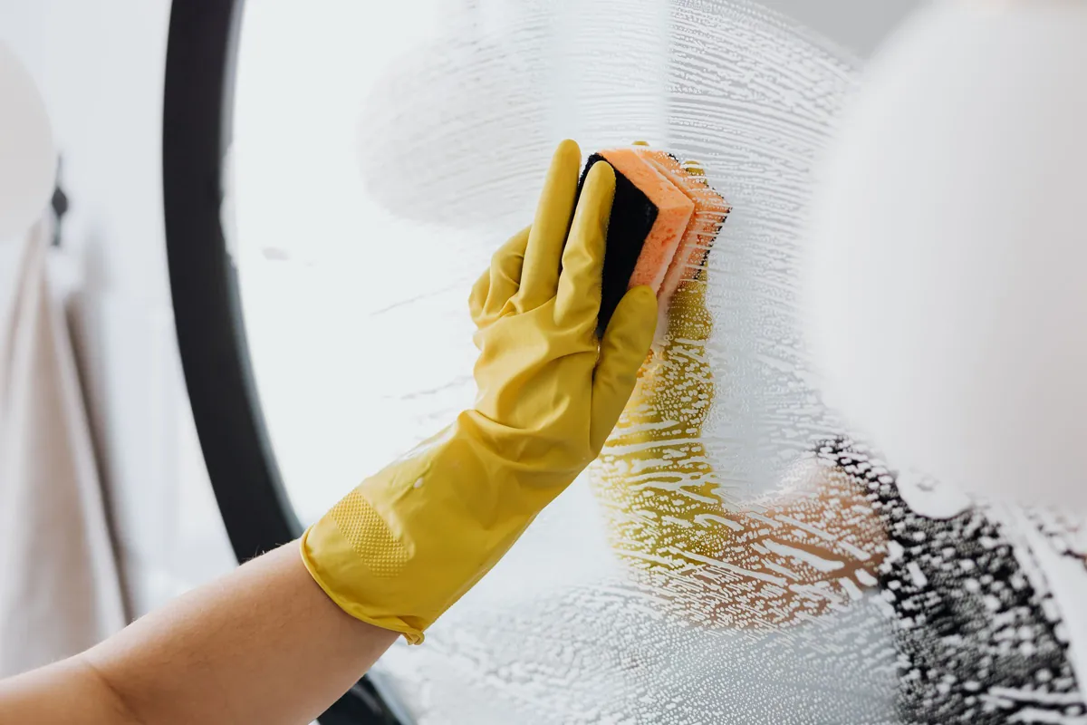A gloved hand washing a mirror with a sponge